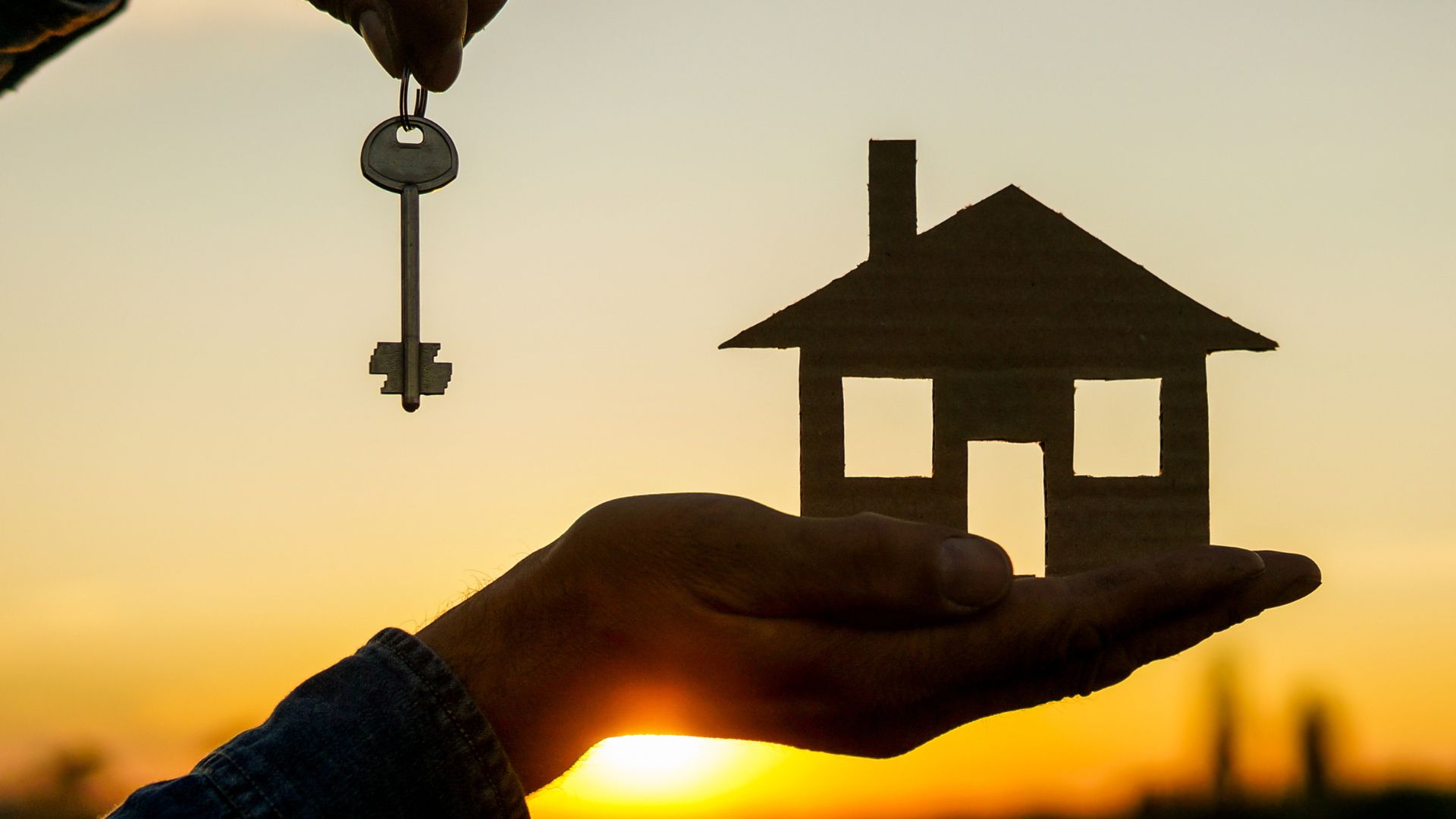 Silhouette of hand holding house cutout and key at sunset